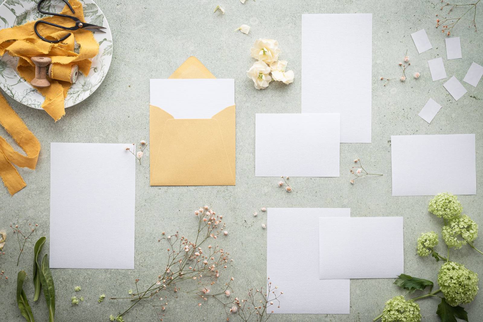 A flat lay of blank stationery and envelope amidst delicate flowers and ribbon, perfect for wedding or event planning.