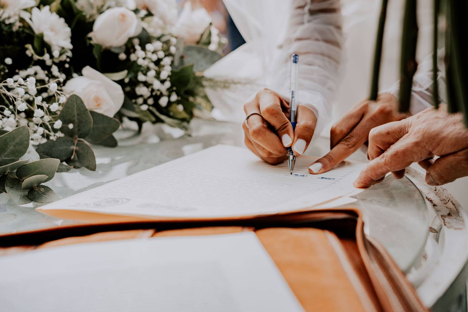 Close-up of a bride signing a wedding certificate with floral arrangement in the background.
