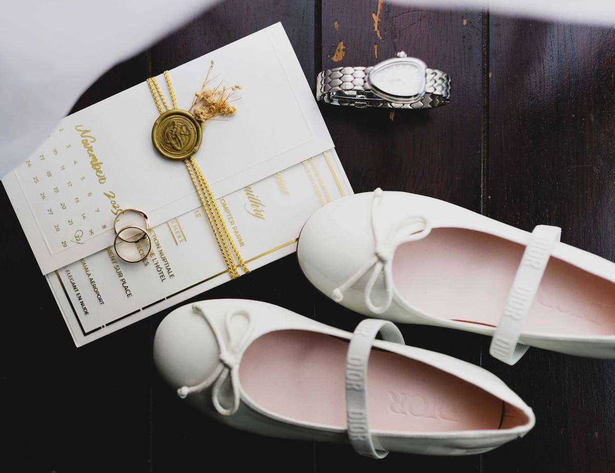 A stylish flat lay of wedding essentials including shoes, invitation, rings, and a watch on dark wood.