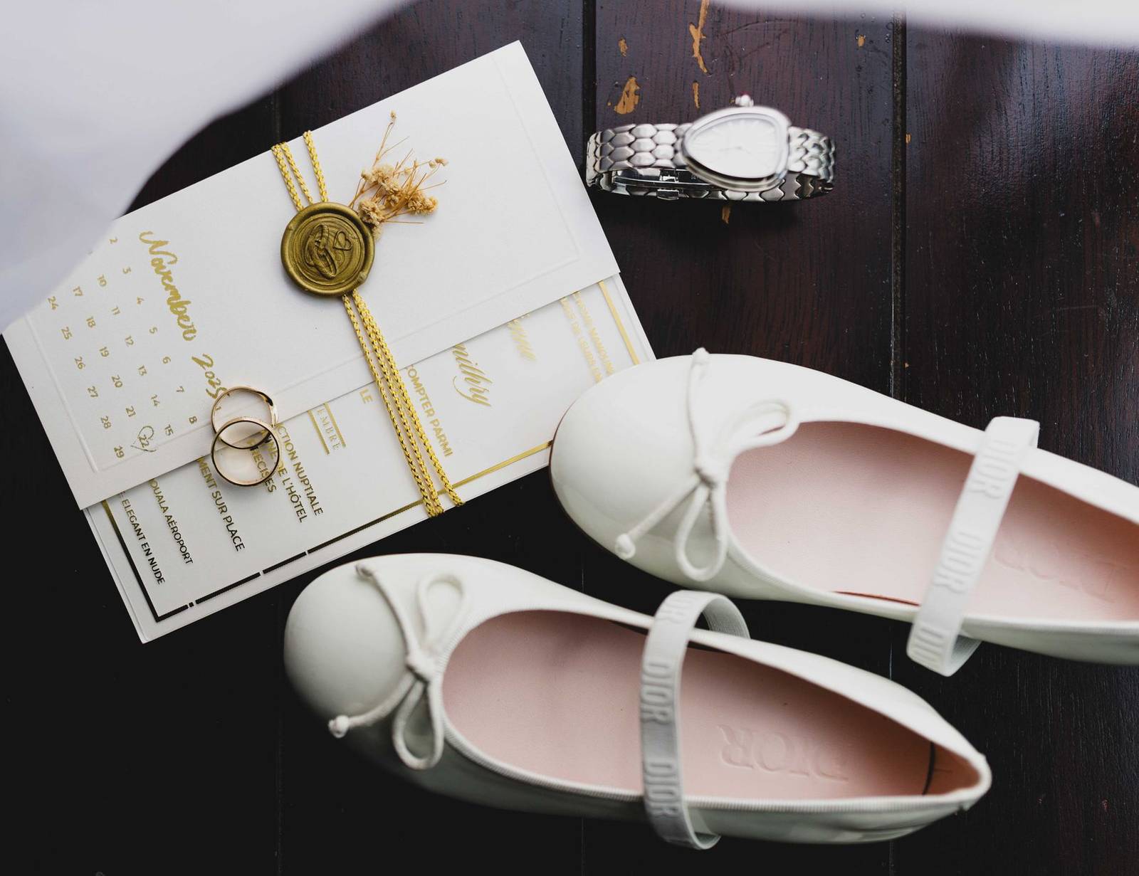 A stylish flat lay of wedding essentials including shoes, invitation, rings, and a watch on dark wood.